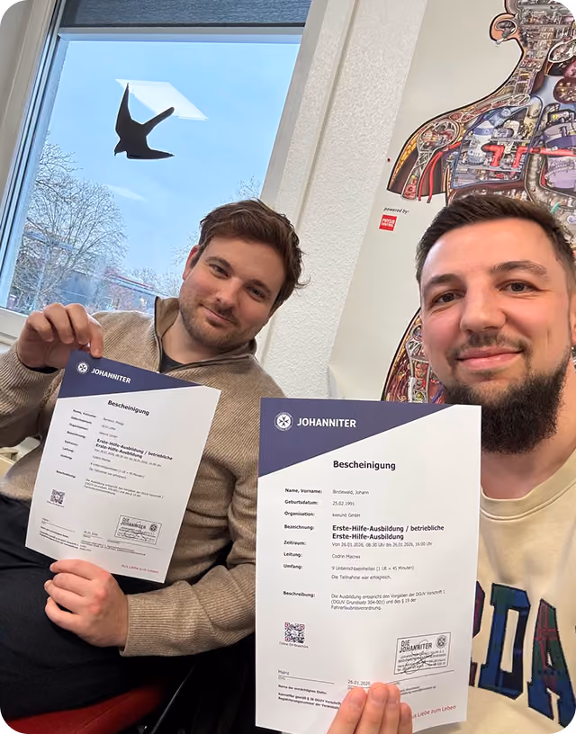 Zwei Männer zeigen Zertifikate ihrer Erste-Hilfe-Ausbildung, hinter ihnen ein Fenster und eine Wandkarte.