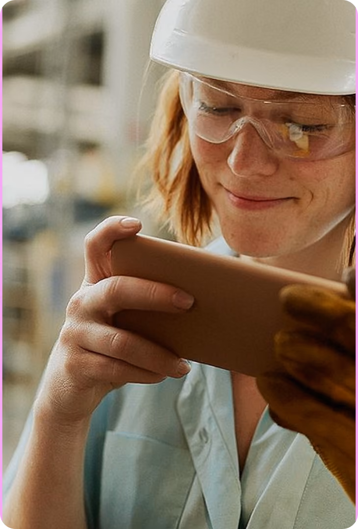 Frau mit Schutzhelm und Brille schaut lächelnd auf ein Gerät in der Hand.