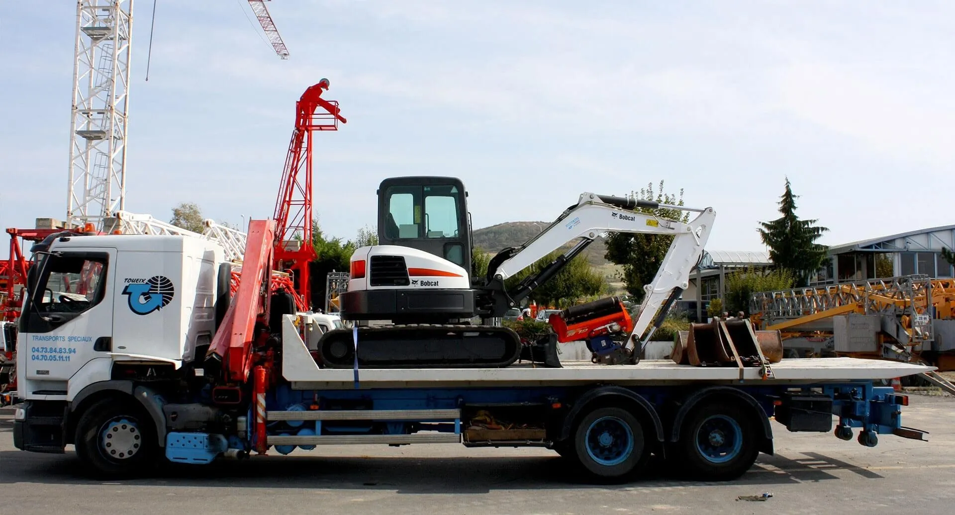 Camion transportant une mini-pelle Bobcat blanche et noire sur une plate-forme, dans un chantier avec des grues et des équipements.