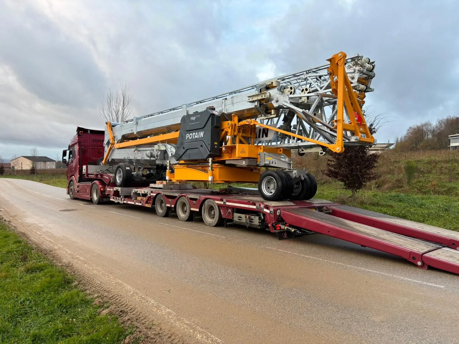 Camion rouge transportant une grue mobile Potain Hup 40 sur une remorque à plusieurs essieux sur une route de campagne.