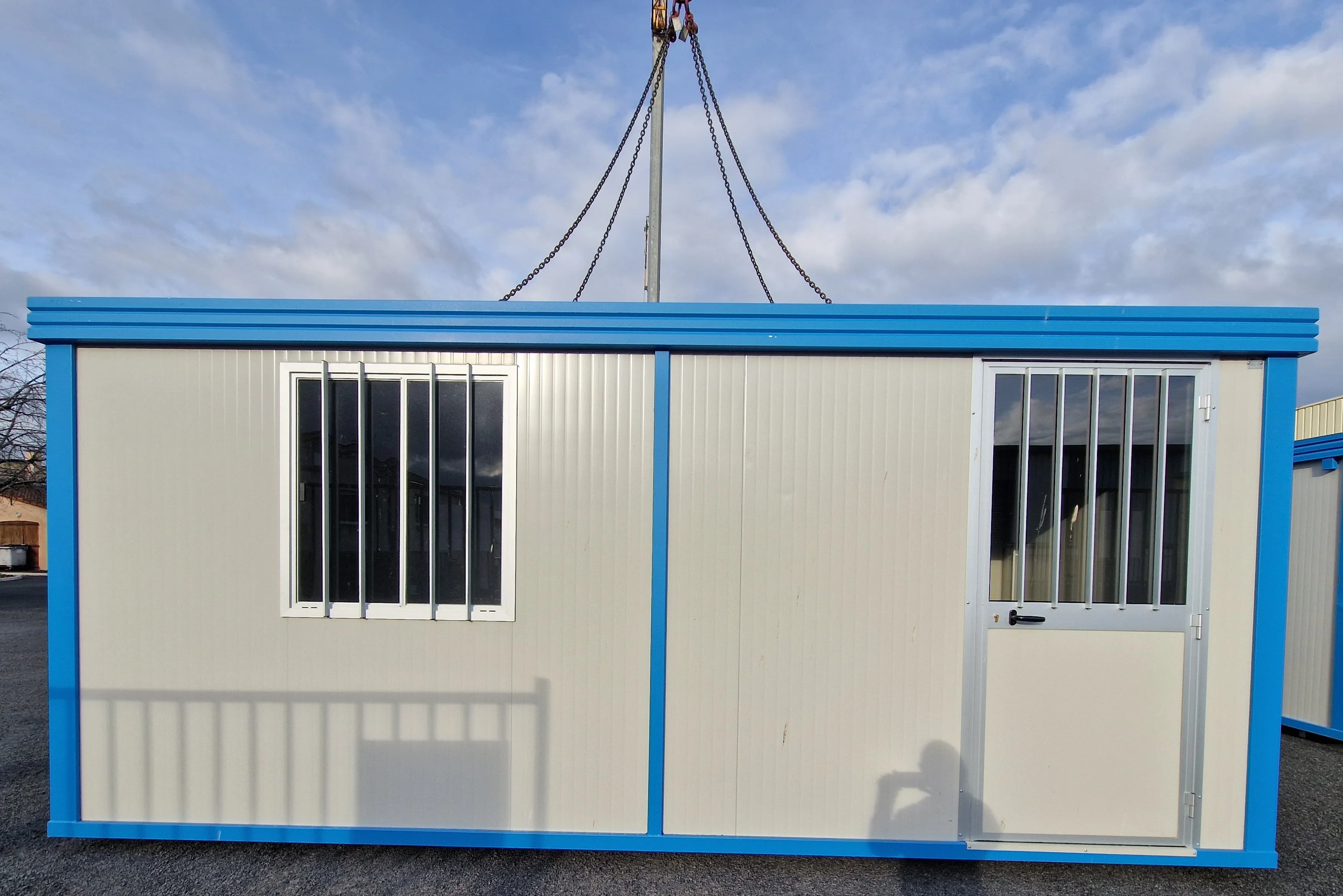 Container modulaire beige avec encadrements bleus, une fenêtre et une porte équipées de grilles métalliques.