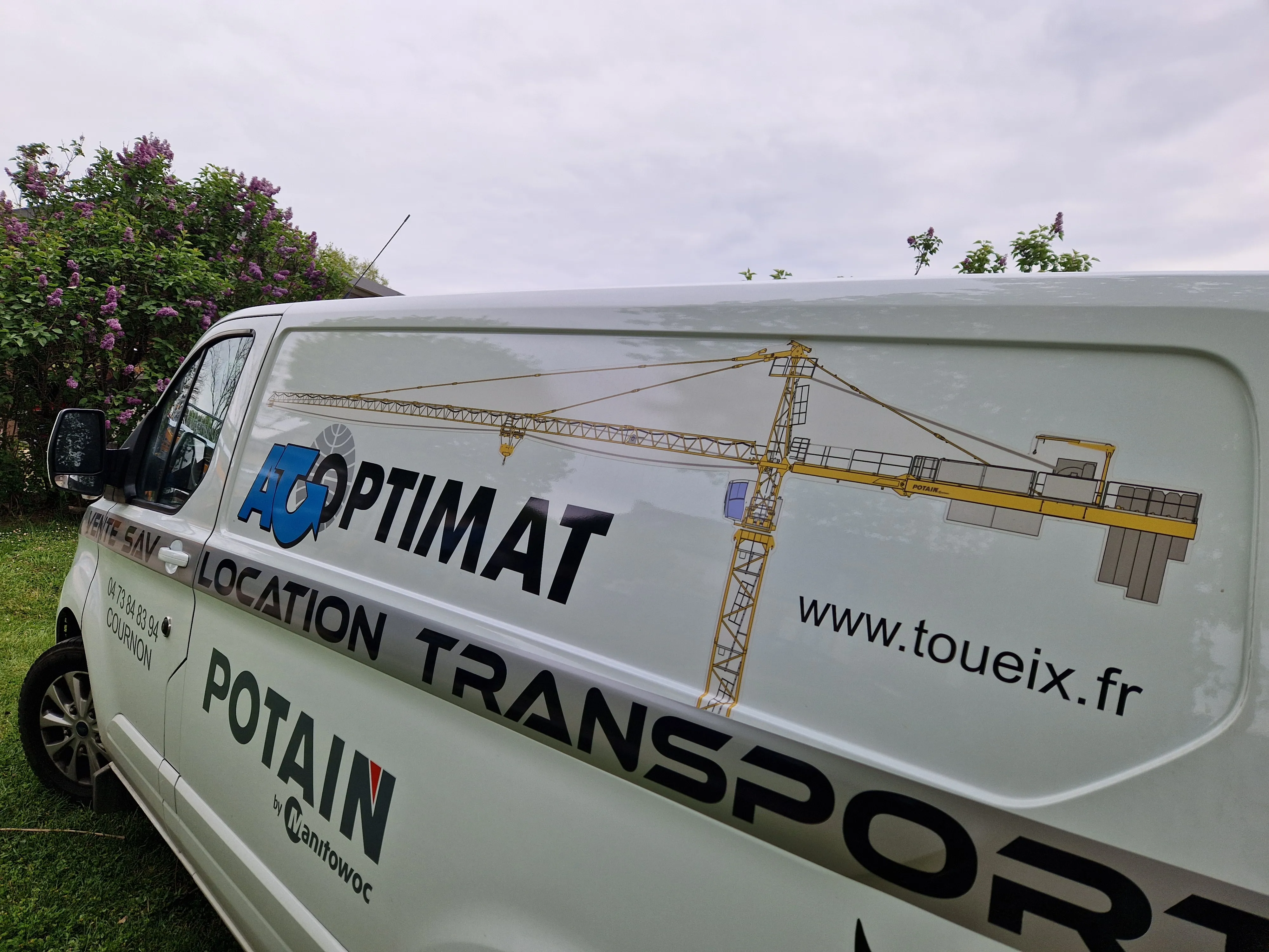 Côté d'une camionnette blanche avec logos et textes publicitaires pour AT OPTIMAT, location transport, POTAIN par Manitowoc, et URL www.toueix.fr, avec illustration d'une grue de construction.