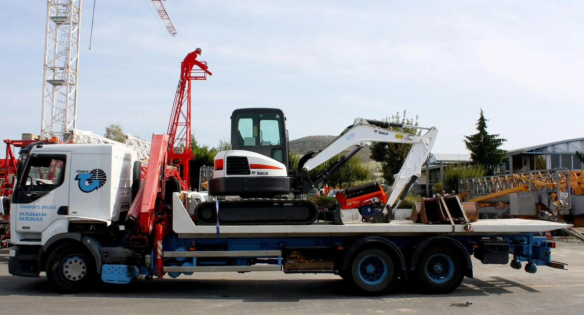 Camion plateau transportant une pelleteuse compacte Bobcat sur un site industriel avec des grues en arrière-plan.