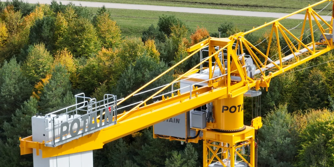 Gros plan d'une grue de construction jaune Potain avec une forêt d'arbres verts et dorés en arrière-plan.