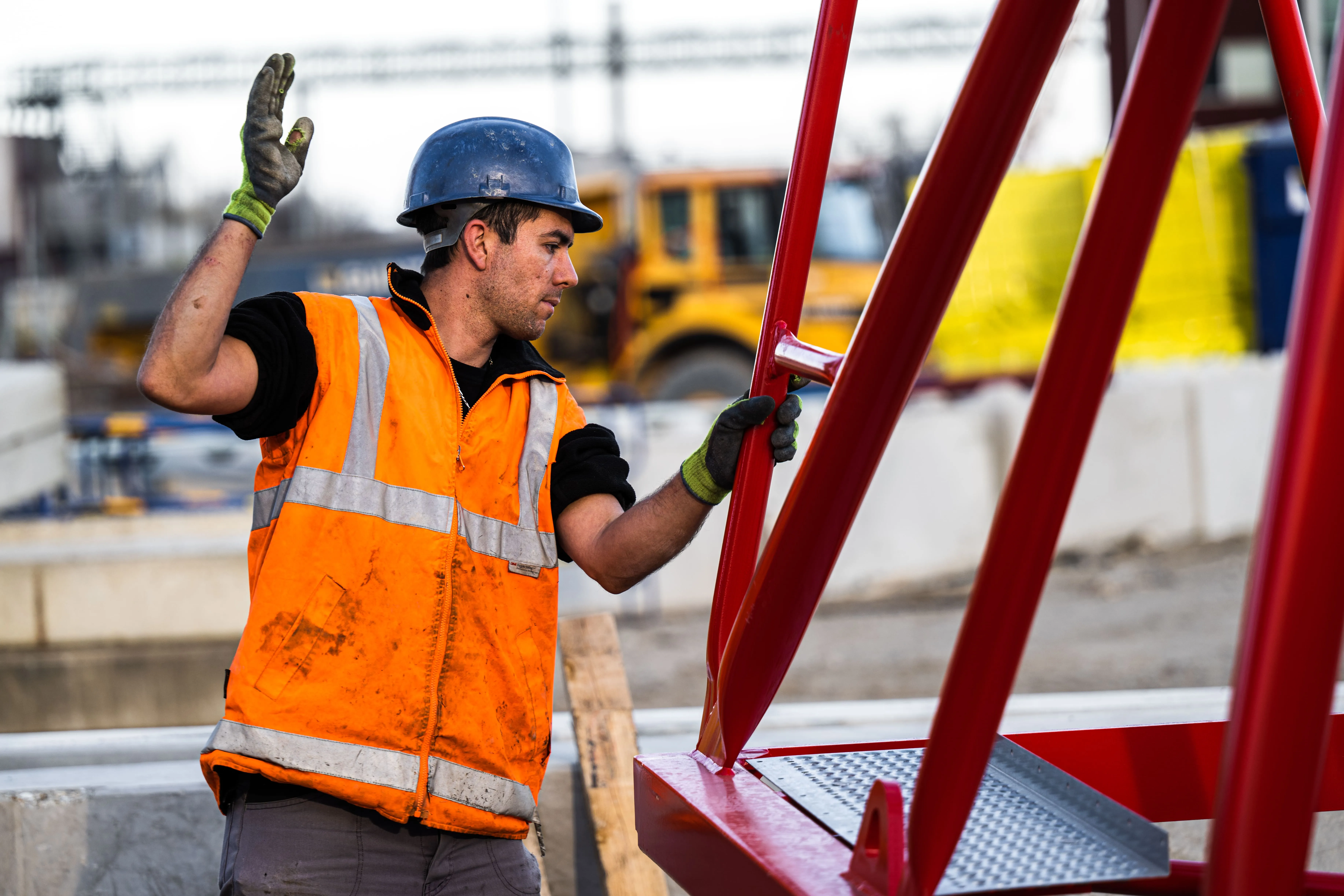 Ouvrier du bâtiment en casque bleu et gilet orange réfléchissant manipulant une grande structure métallique rouge sur un chantier.