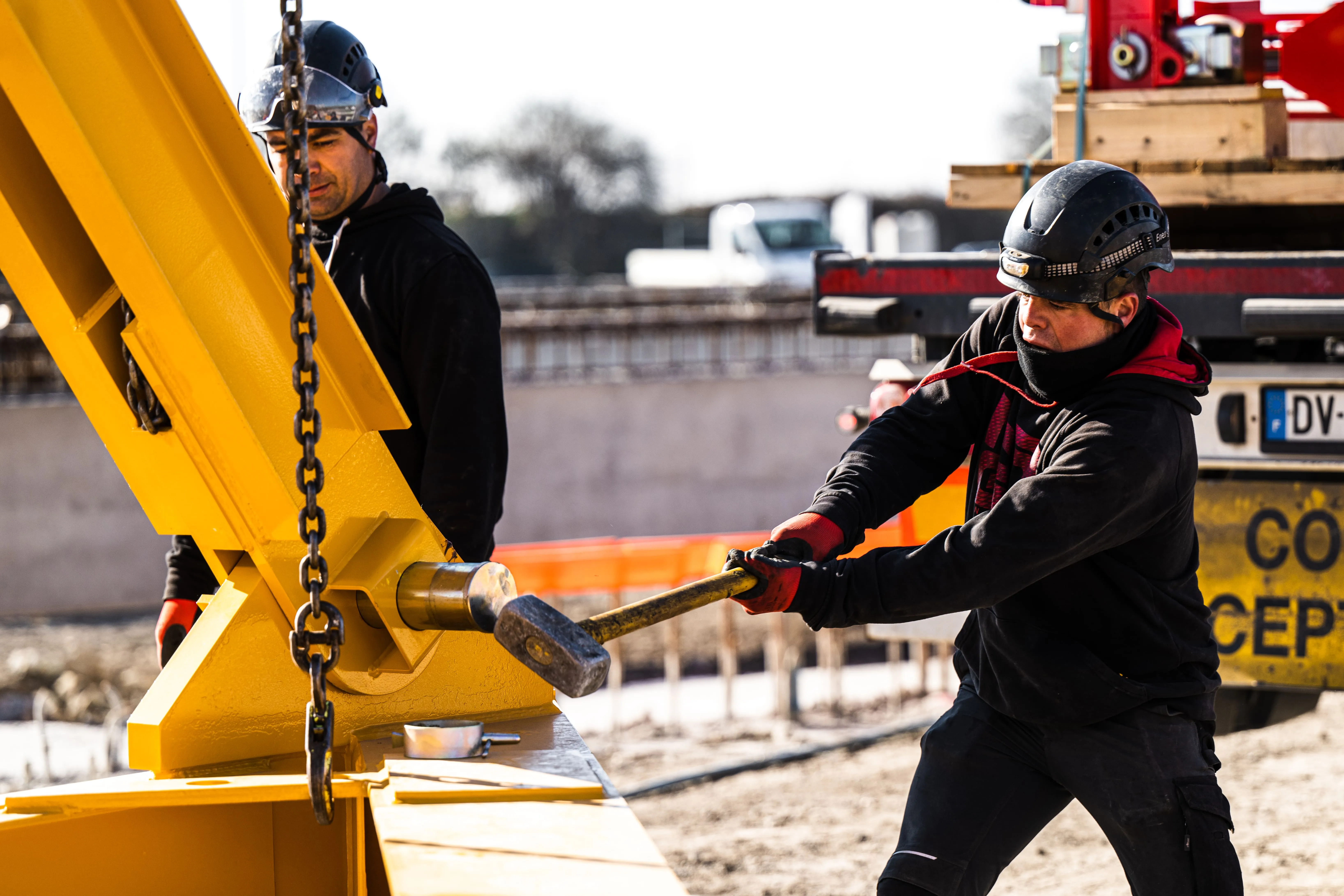 Deux ouvriers avec casques de sécurité travaillent sur une grande structure métallique jaune sur un chantier.