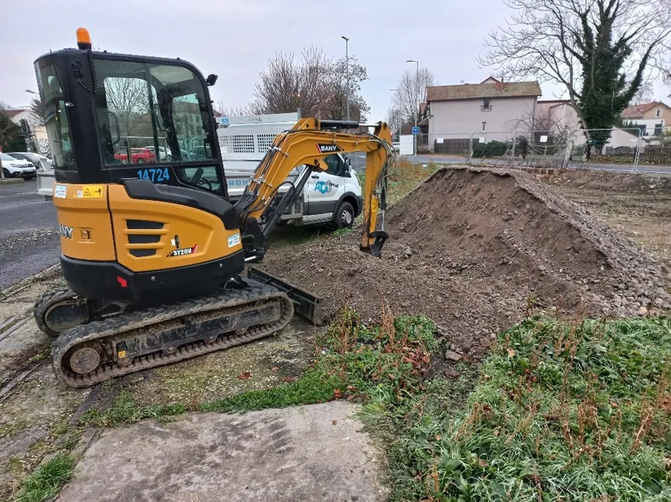 Mini pelle jaune avec chenilles sur un chantier à côté d'un tas de terre et d'une camionnette blanche.