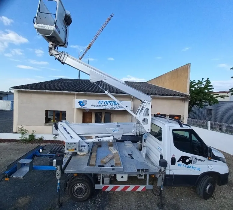 Camion utilitaire blanc équipé d'une nacelle élévatrice déployée sur un site extérieur devant un bâtiment industriel.
