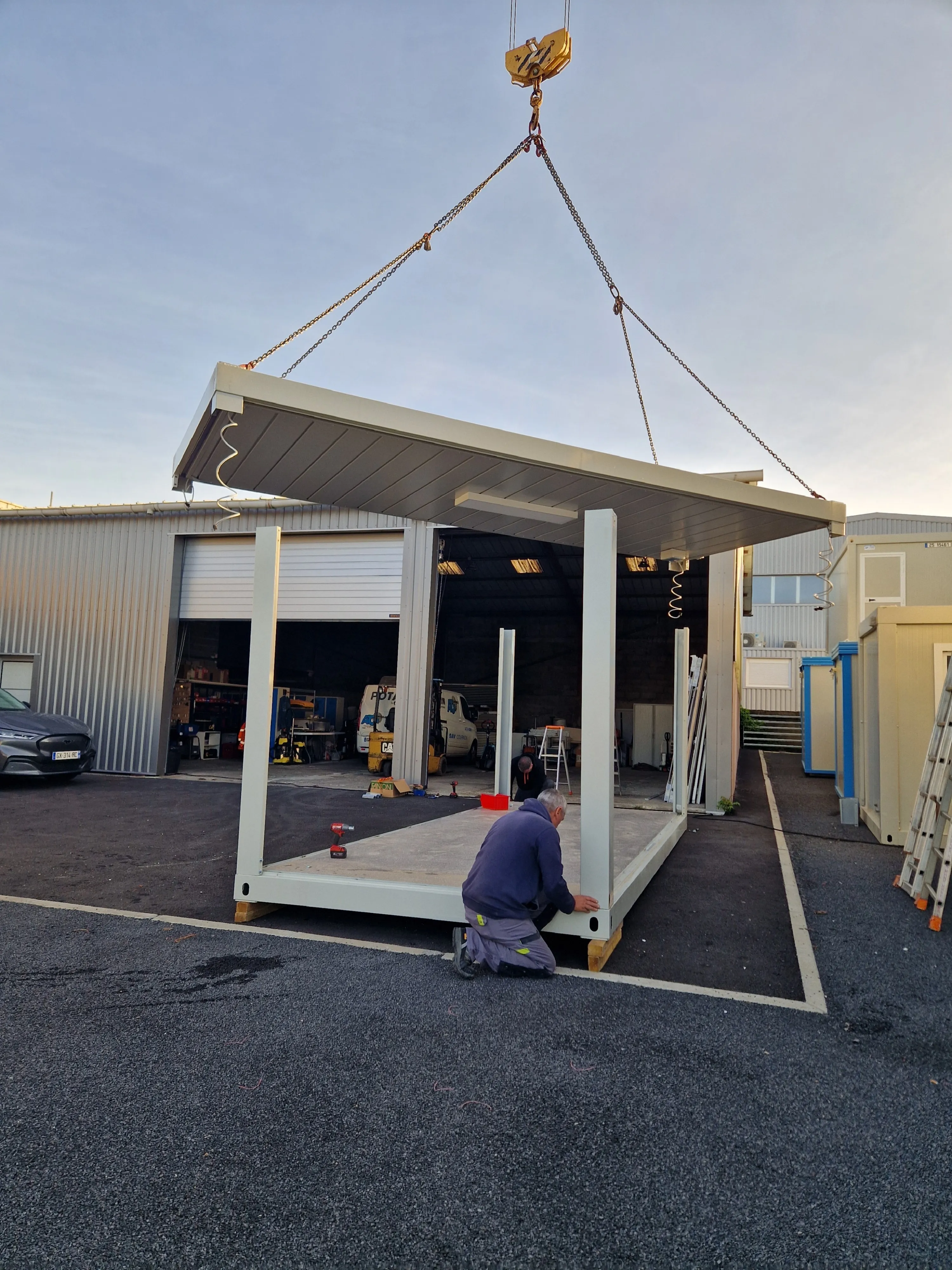 Deux hommes assemblent une structure modulaire tandis qu'un panneau de toit est suspendu par une grue.