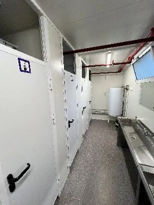 Intérieur d'une toilette publique avec cabines blanches et lavabo en inox à droite.