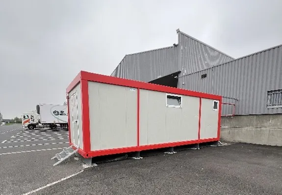 Bâtiment portable gris avec cadre rouge installé sur un parking devant un entrepôt industriel.