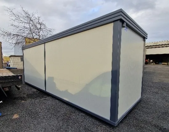 Conteneur modulaire beige à cadres gris en extérieur sur sol gravillonné, sous ciel nuageux.