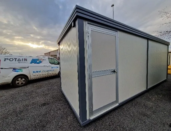 Bâtiment modulaire blanc avec porte en métal encadré gris, camion avec logos en arrière-plan au crépuscule.