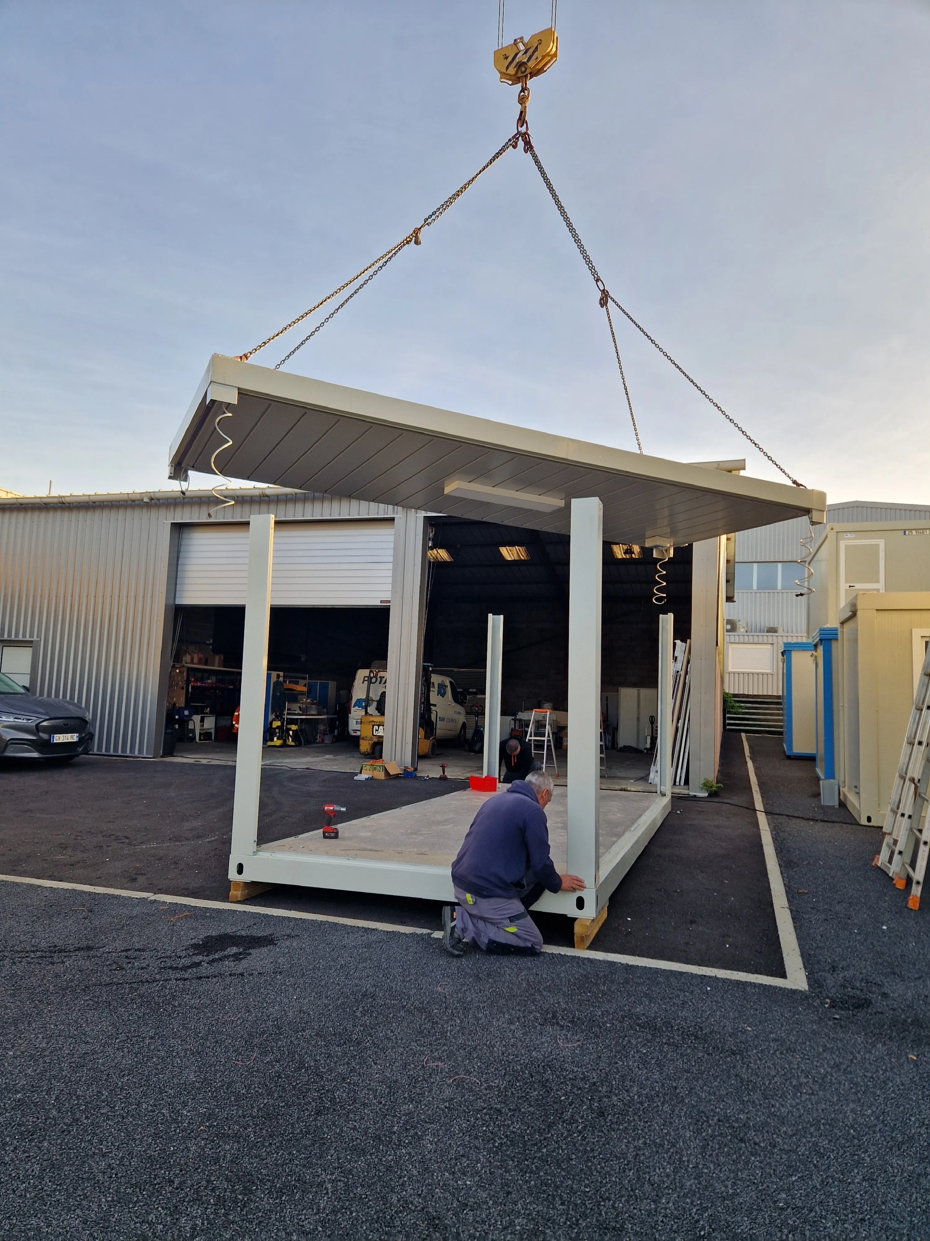 Deux ouvriers assemblent une structure modulaire avec un toit levé par une grue.
