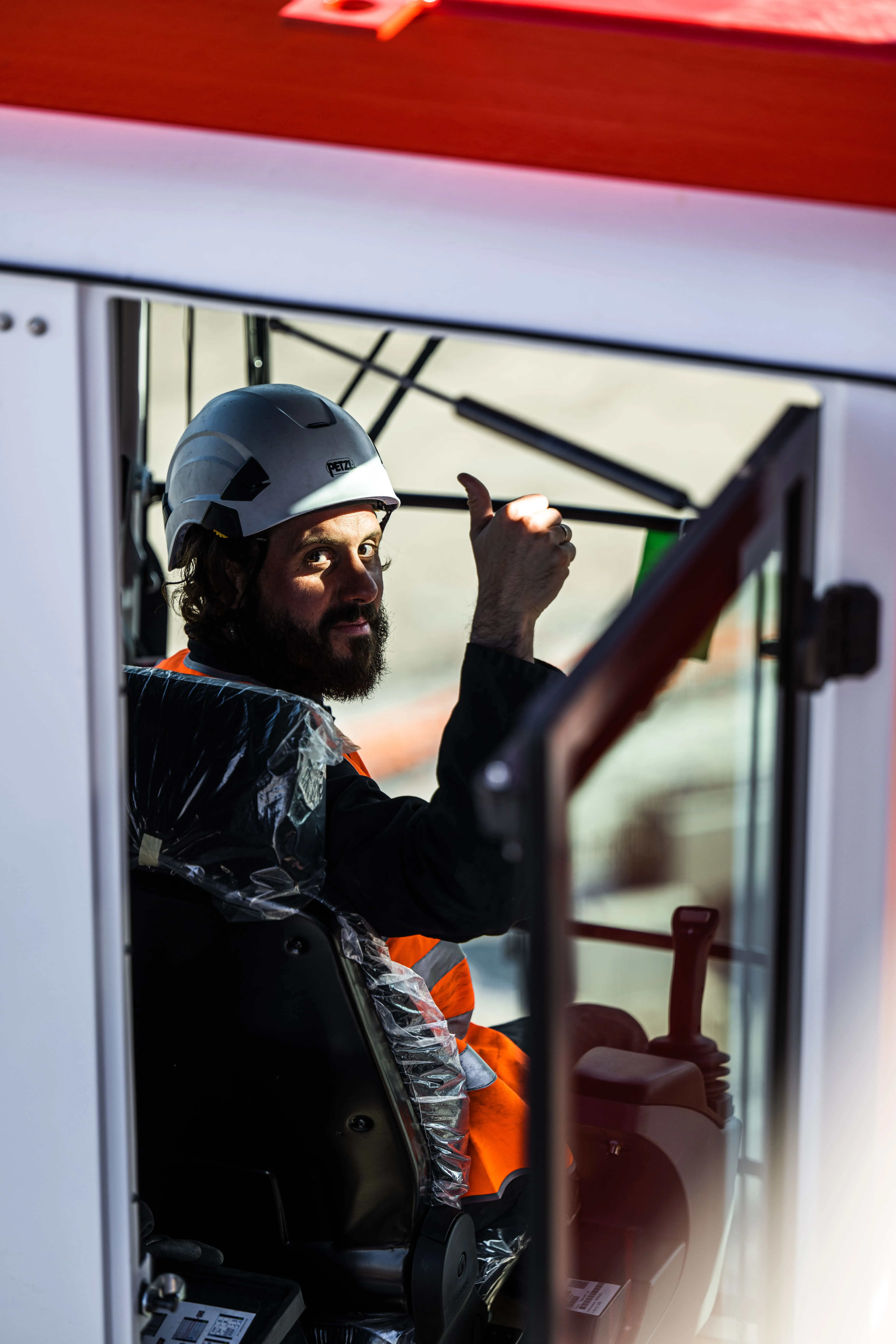 Ouvrier portant un casque et un gilet orange fait un signe pouce levé depuis la cabine d'une grue.