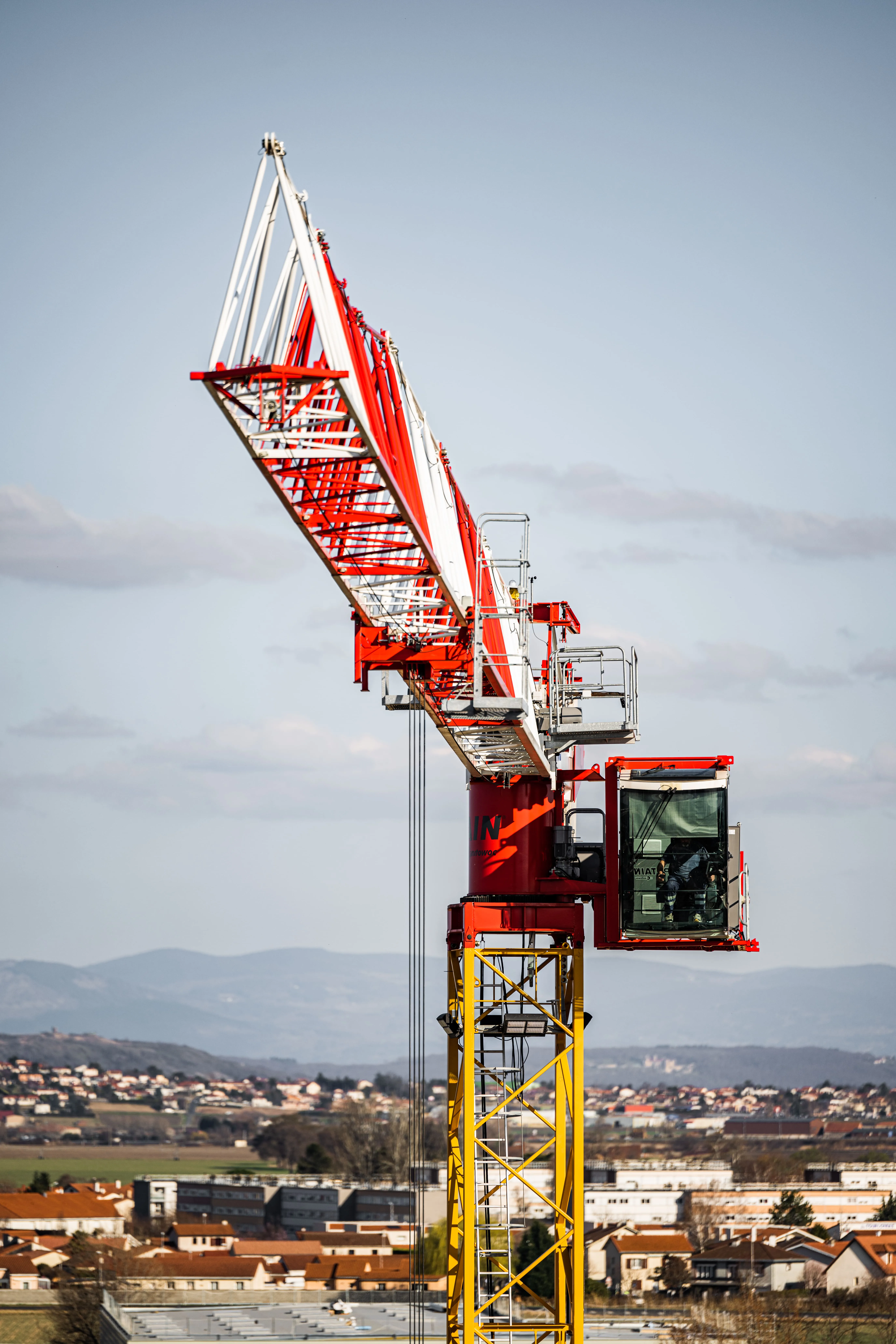Grue de construction rouge et jaune sur un site avec des maisons et des montagnes en arrière-plan.