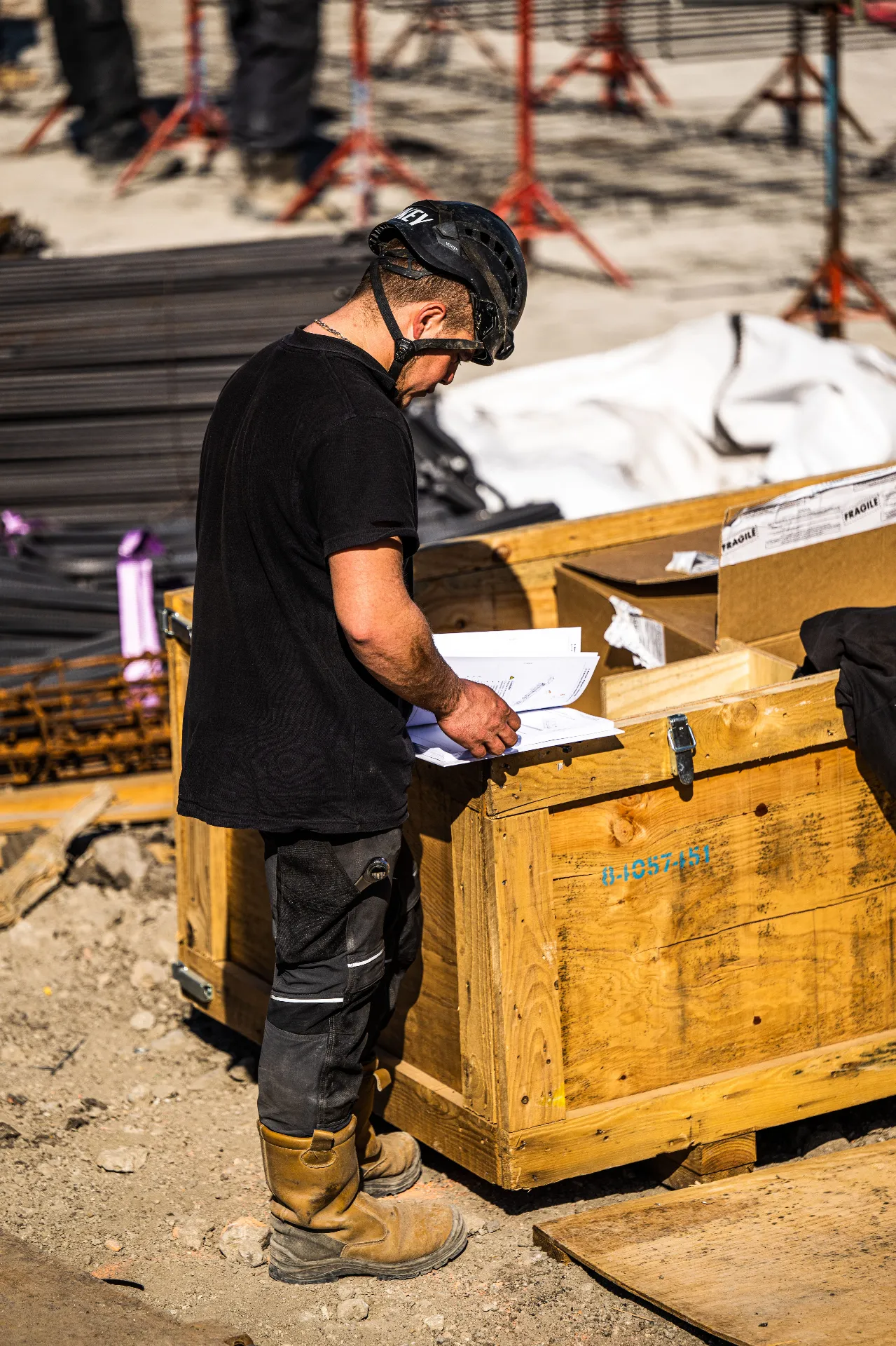Un ouvrier portant un casque consulte des documents sur un chantier à côté d'une caisse en bois.