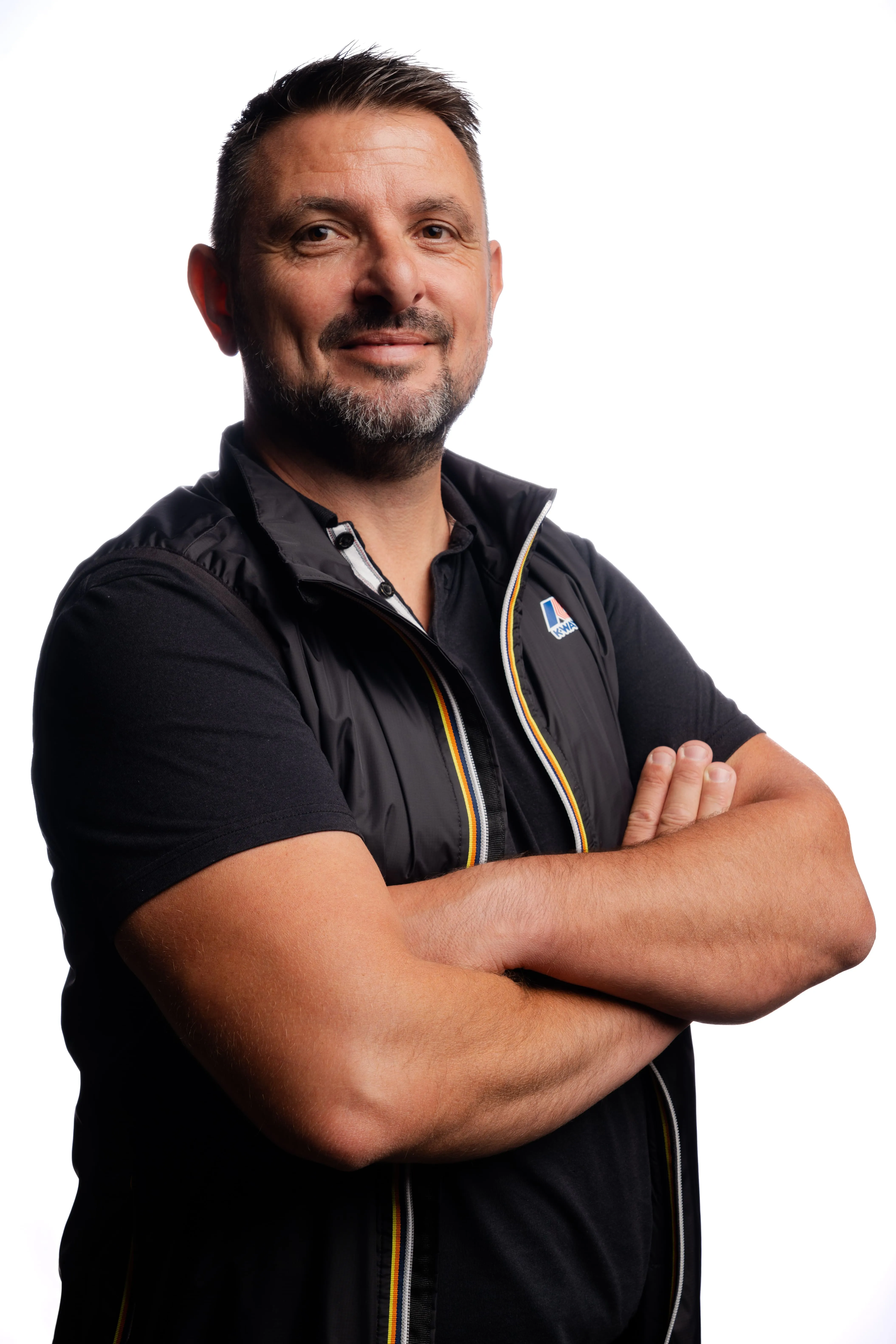 Homme souriant les bras croisés, portant un gilet noir et un t-shirt noir sur fond blanc.