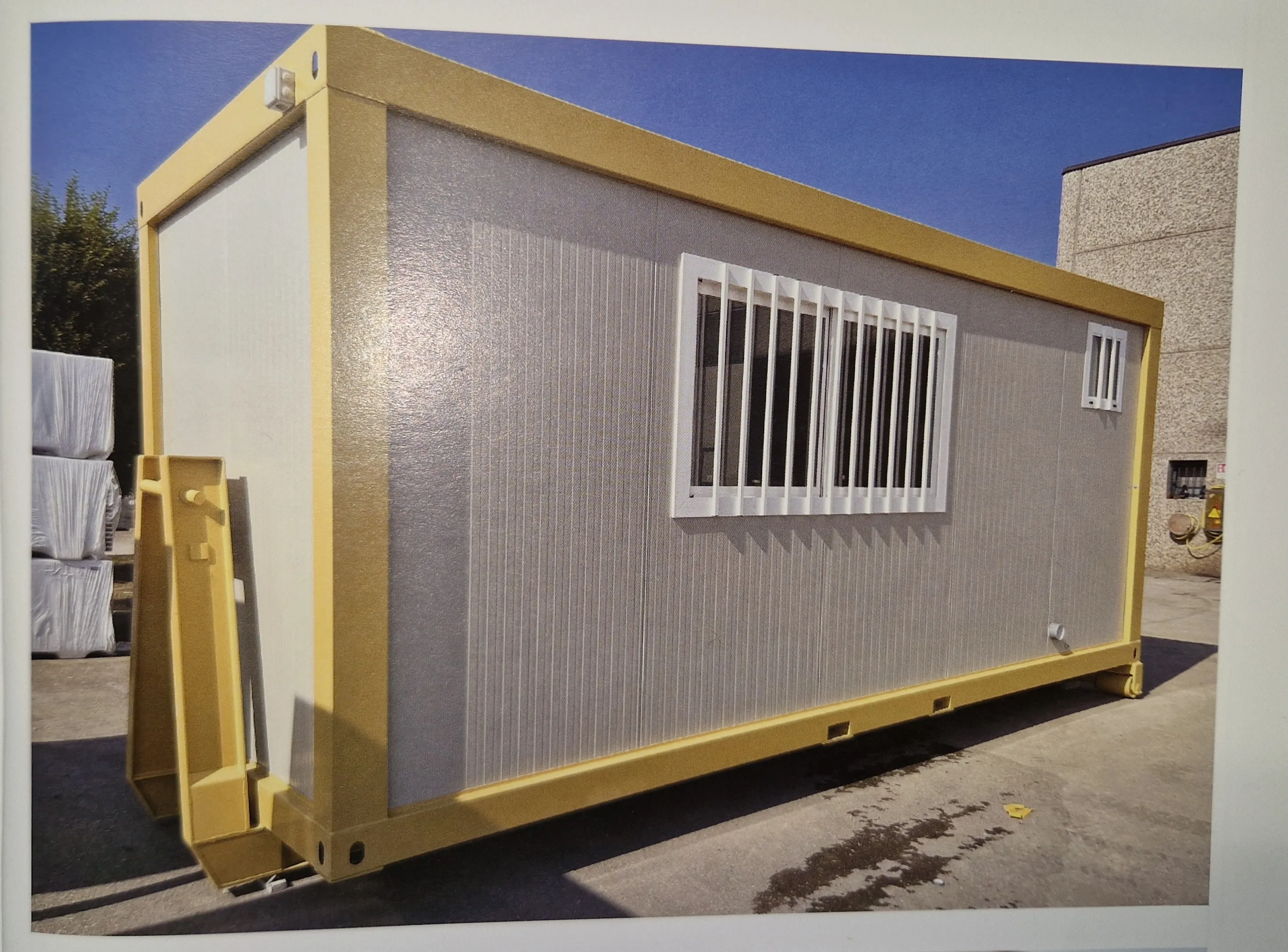 Conteneur modulaire gris avec cadre jaune et fenêtre avec barreaux blancs.