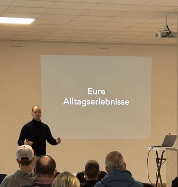 Ein Mann in schwarzem Pullover hält einen Vortrag vor sitzendem Publikum, auf der Wand hinter ihm steht ‚Eure Alltagserlebnisse‘.