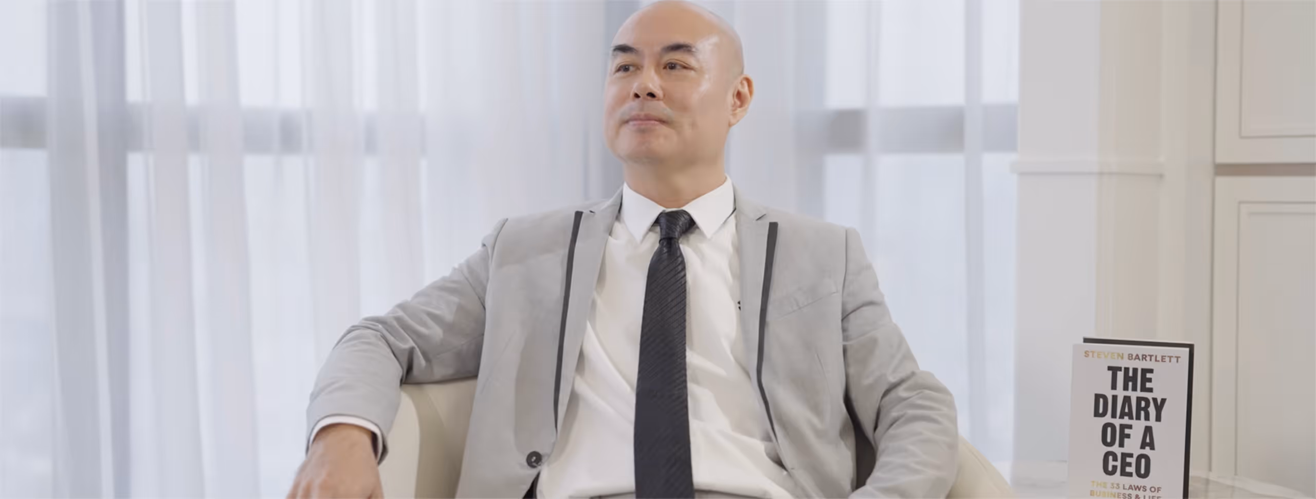 Ein Mann in grauem Anzug und schwarzer Krawatte sitzt entspannt auf einem weißen Stuhl, daneben Buch 'The Diary of a CEO'.