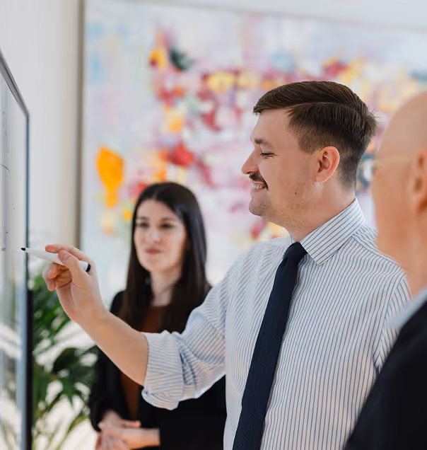 Geschäftsmann schreibt an ein Whiteboard, während zwei Kollegen im Hintergrund zuhören.