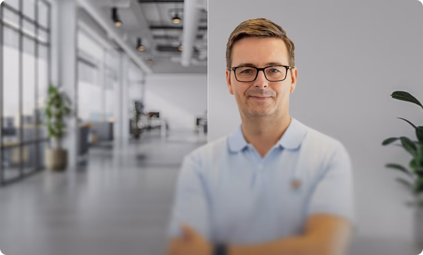 Porträt eines Mannes mit Brille und hellblauem Poloshirt vor verschwommenem Bürohintergrund.