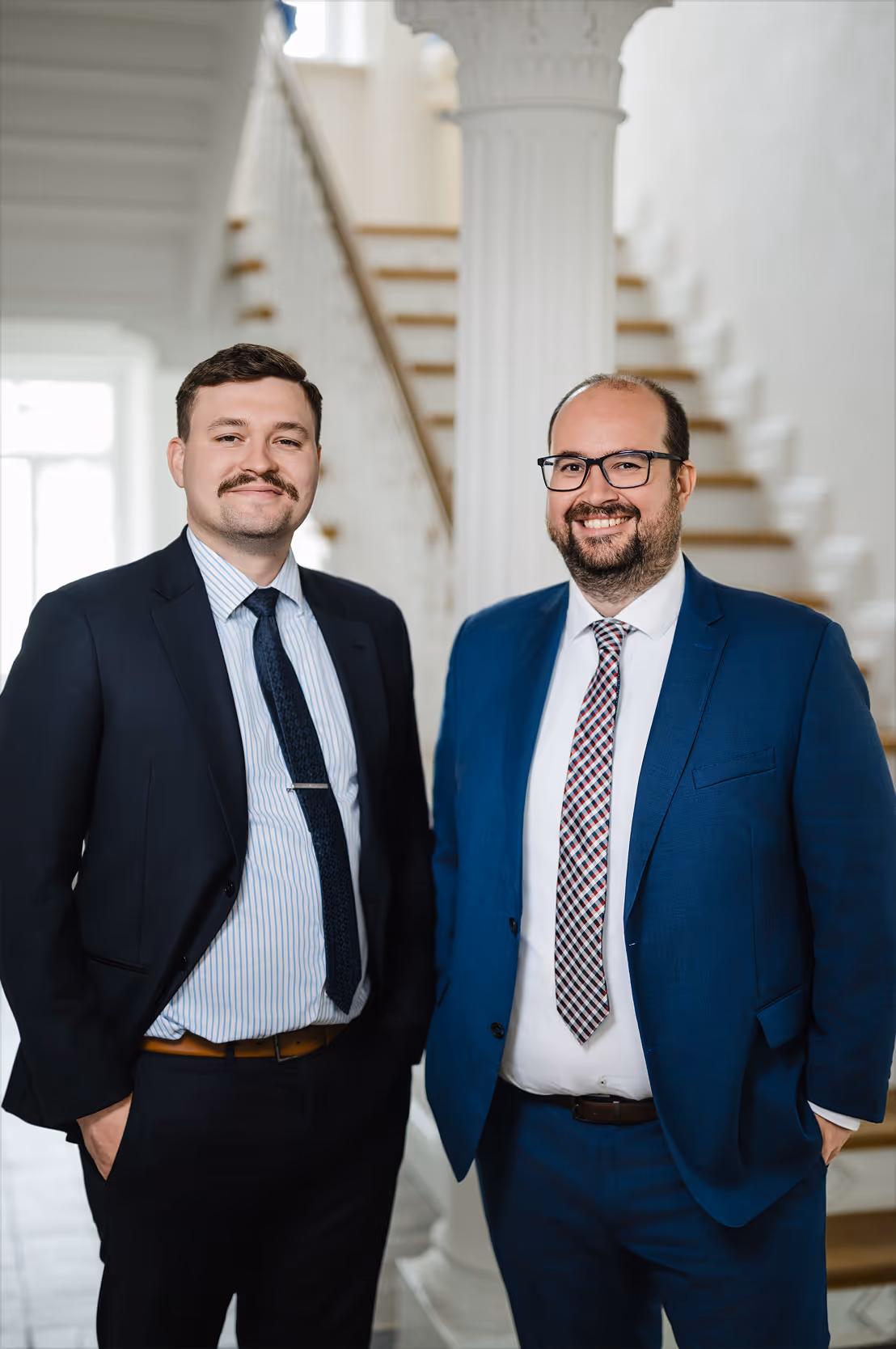 Zwei Männer in Anzügen stehen lächelnd nebeneinander in einem Innenraum mit Treppe und Säule im Hintergrund.