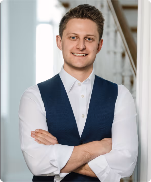 Junger Mann mit weißem Hemd und blauem Weste lächelt mit verschränkten Armen vor einer hellen Treppe.
