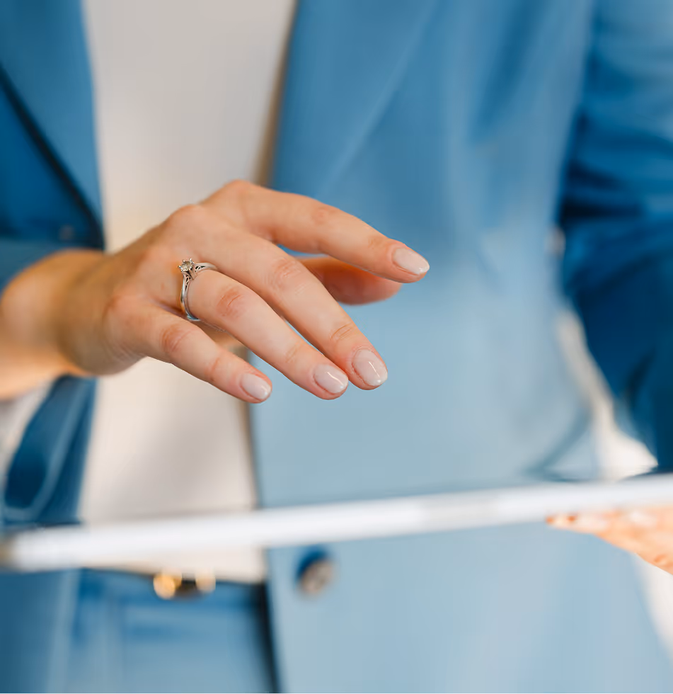 Nahaufnahme einer Hand mit Fingerring, die auf ein Tablet zeigt, im Hintergrund eine Person mit blauem Anzug.
