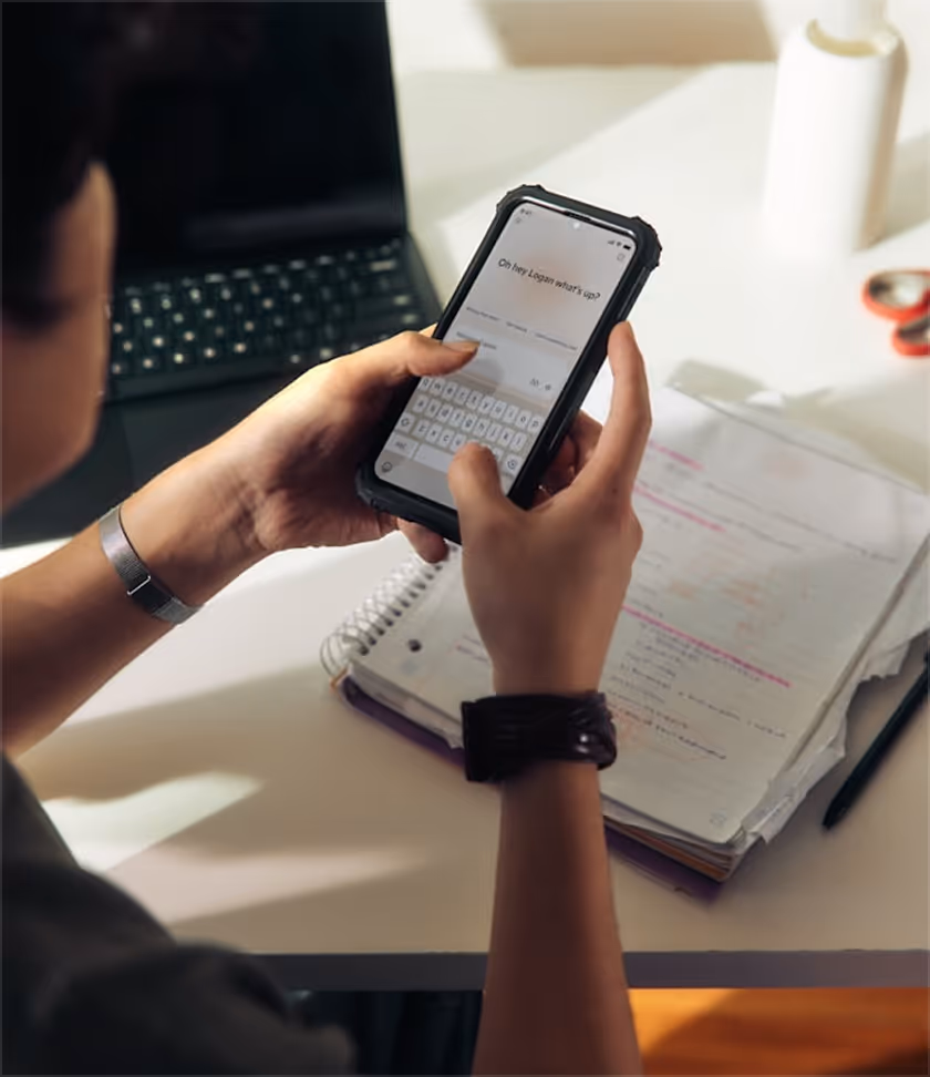 Person tippt eine Nachricht auf einem Smartphone, neben Notizblock, Stift und Laptop auf einem Schreibtisch.