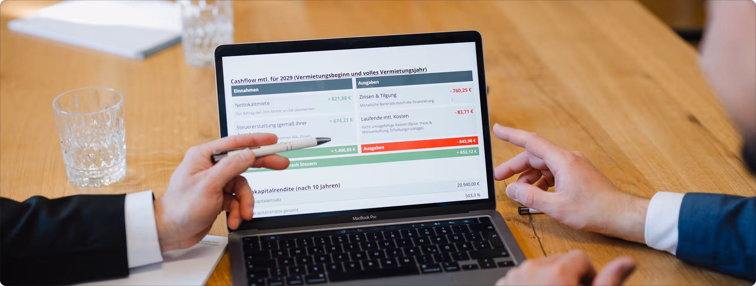 Zwei Personen diskutieren über monatlichen Cashflow und Kapitalrendite auf einem MacBook Pro, mit Finanzzahlen und Ausgabenübersicht auf dem Bildschirm.