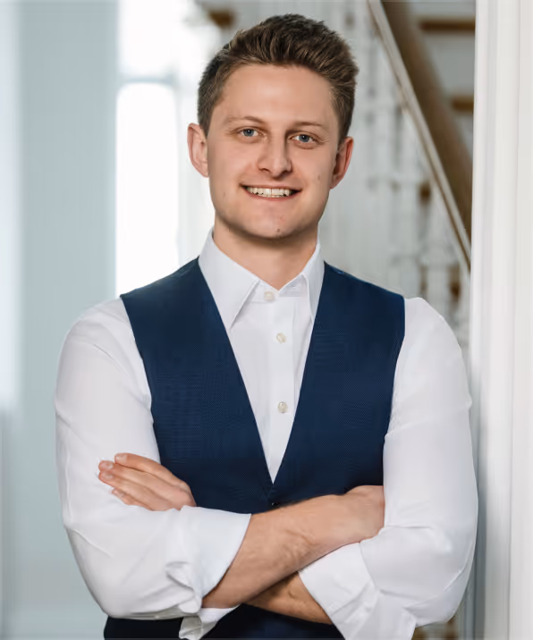 Junger Mann mit weißen Hemdärmeln und blauem Weste, der lächelnd die Arme vor der Brust verschränkt.