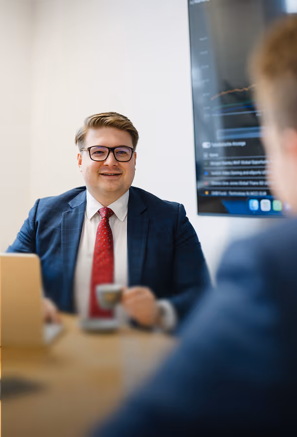 Lächelnder Mann in dunklem Anzug und roter Krawatte sitzt beim Meeting mit Kaffee und Laptop vor einem Bildschirm mit Diagrammen.
