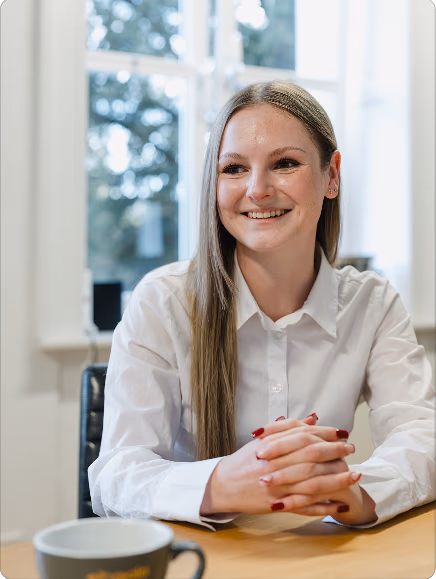 Junge Frau mit langen blonden Haaren und weißem Hemd, die lächelnd an einem Tisch sitzt, vor einem Fenster.