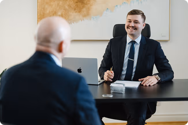 Zwei Männer im Anzug führen ein professionelles Gespräch in einem Büro, einer sitzt am Schreibtisch mit einem Laptop und Notizen.