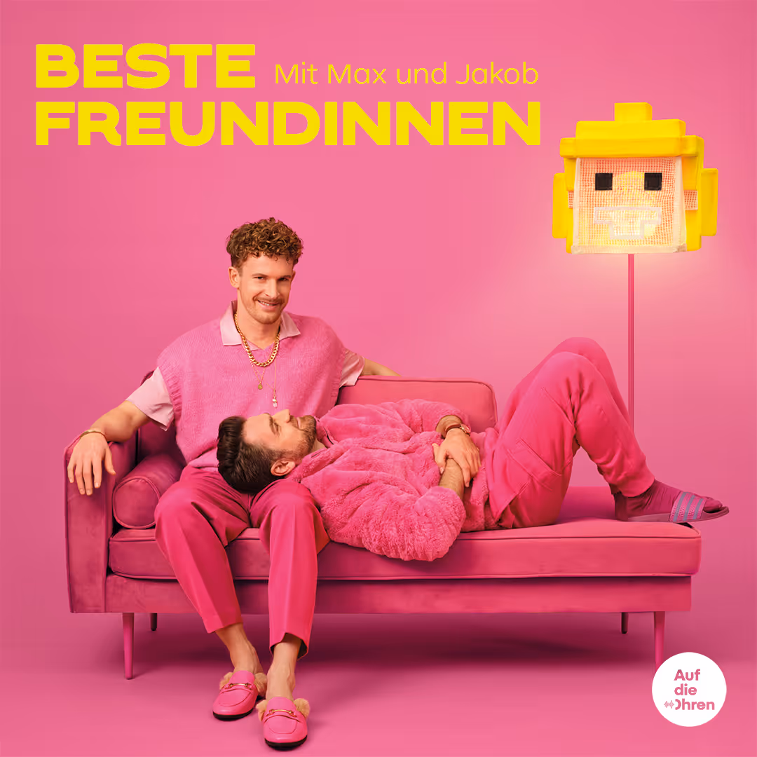 Zwei Männer, einer liegt auf pinkem Sofa, der andere sitzt, alle in Pink mit gelber Lampe im Raum.