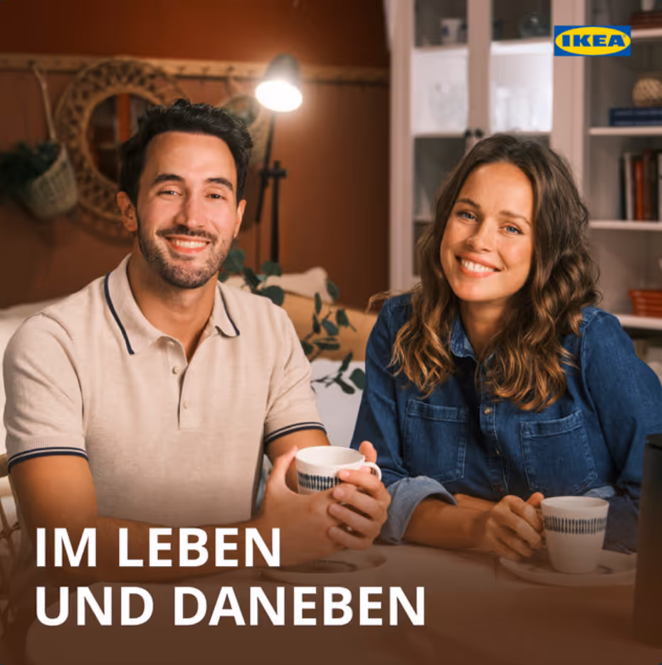 Lächelndes Paar sitzt an Tisch mit Tassen vor warmer Wohnzimmerkulisse.