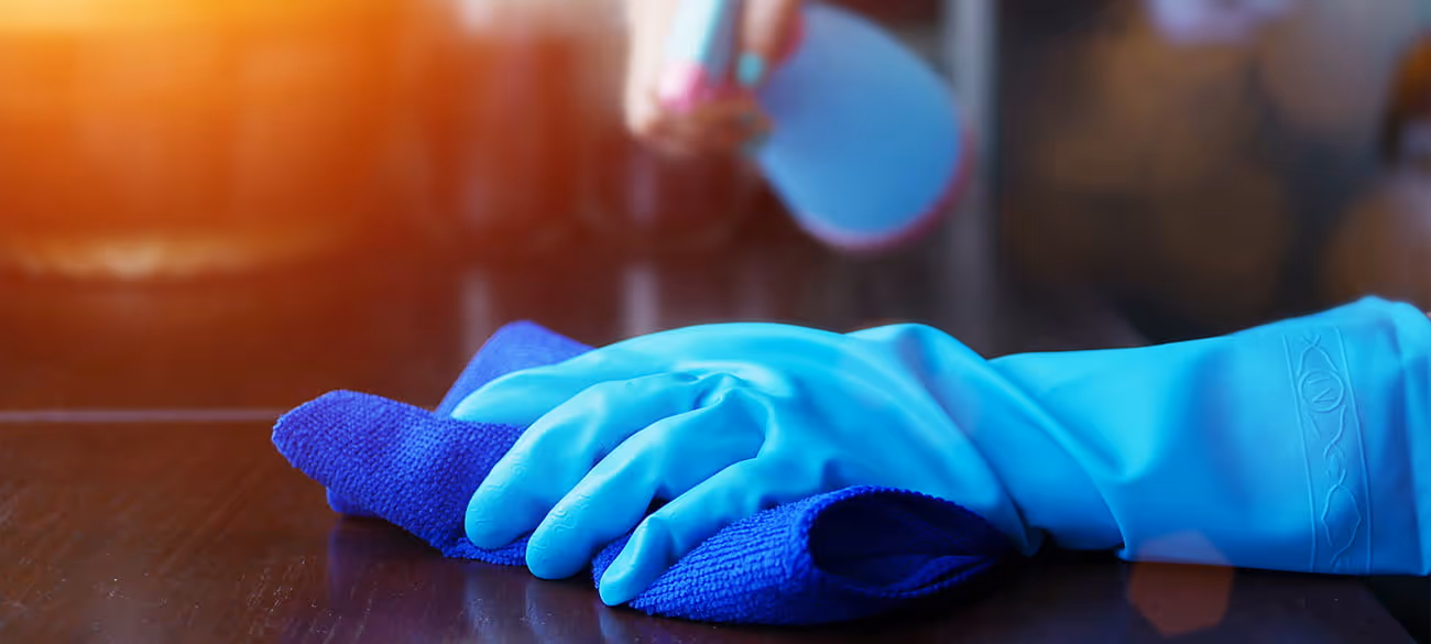 Hand mit blauen Gummihandschuhen wischt eine dunkle Holzoberfläche mit einem blauen Reinigungstuch.