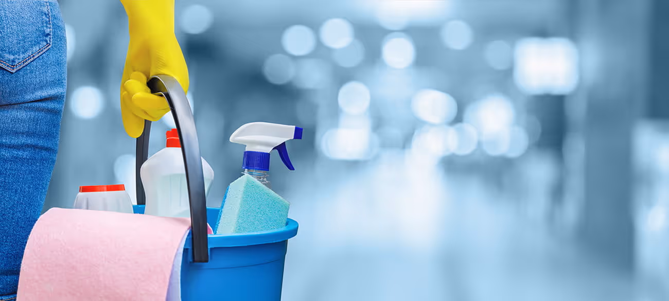 Person mit gelben Handschuhen hält blauen Eimer mit Reinigungsmitteln und rosa Tuch vor verschwommenem Hintergrund.