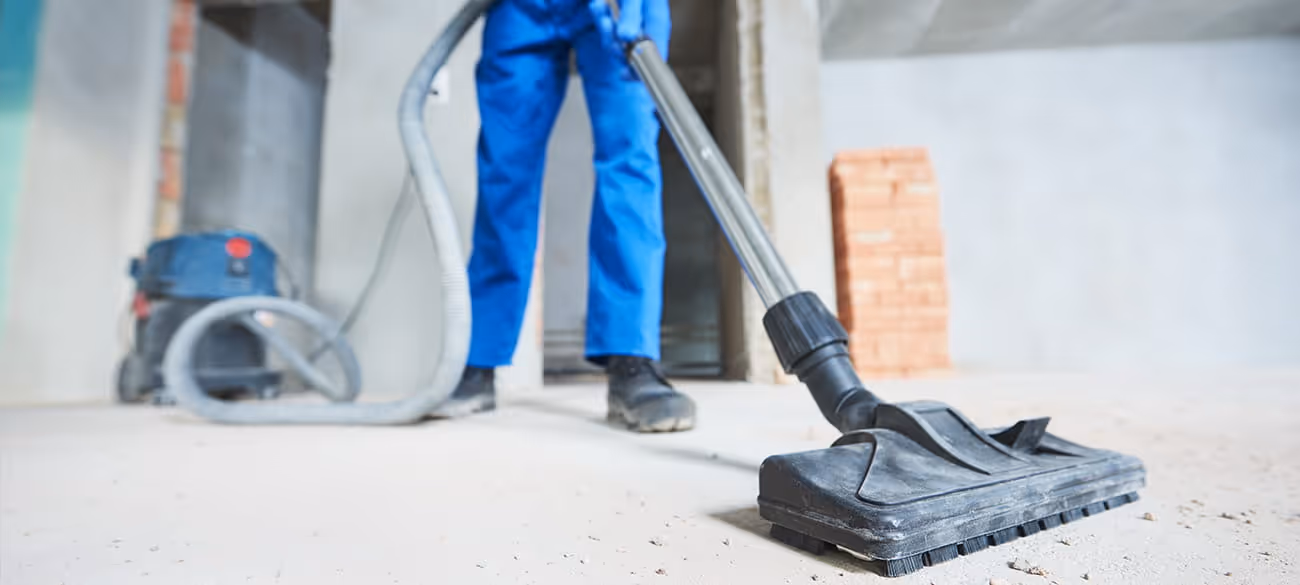 Person in blauer Arbeitskleidung, die eine Baustelle mit einem Industriesauger reinigt.