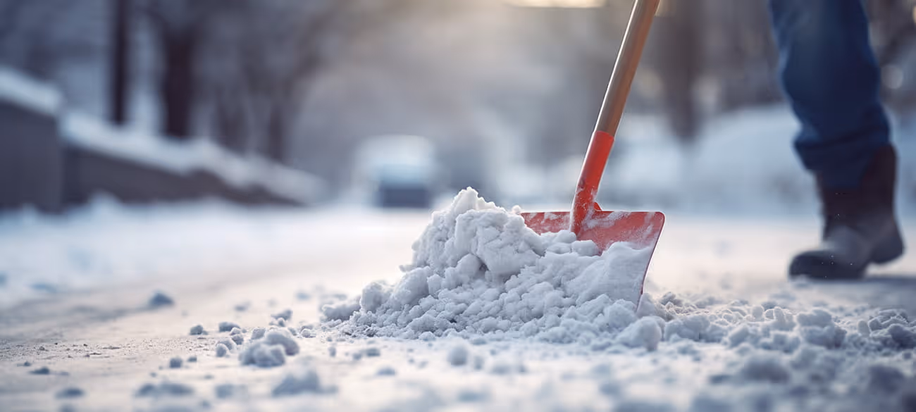 Person mit Stiefeln schiebt Schnee mit einer roten Schneeschaufel auf einem verschneiten Weg.