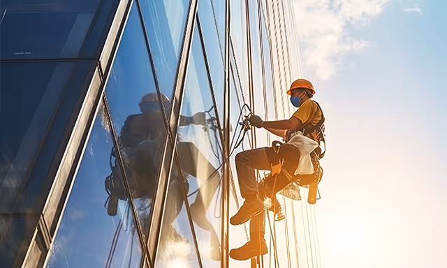 Fensterputzer mit Schutzhelm und Maske reinigt die Fassade eines hohen Bürogebäudes.
