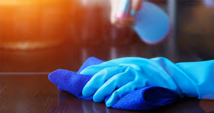 Hand mit blauem Handschuh reinigt Oberfläche mit blauem Tuch und Sprühflasche im Hintergrund.