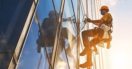 Fensterputzer in Schutzkleidung reinigt Hochhausfenster von außen im Sonnenlicht.