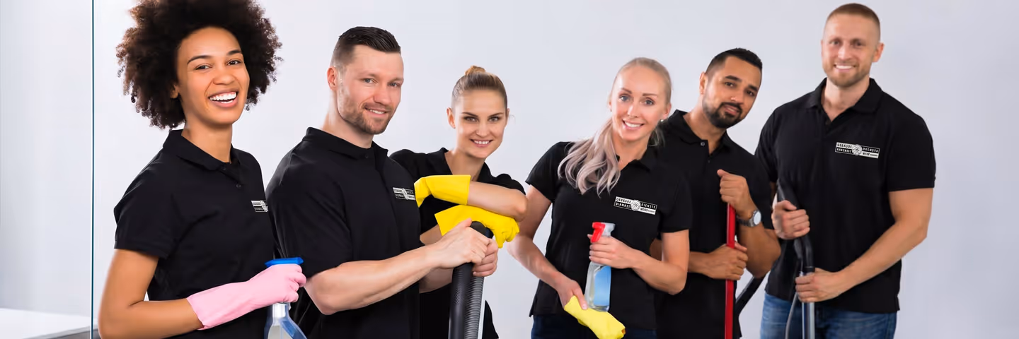 Sechs Reinigungskräfte in schwarzen Poloshirts mit Handschuhen und Putzutensilien lächeln in die Kamera.