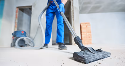 Arbeiter in blauer Arbeitskleidung saugt mit Industriesauger auf Baustelle Staub vom Boden ab.