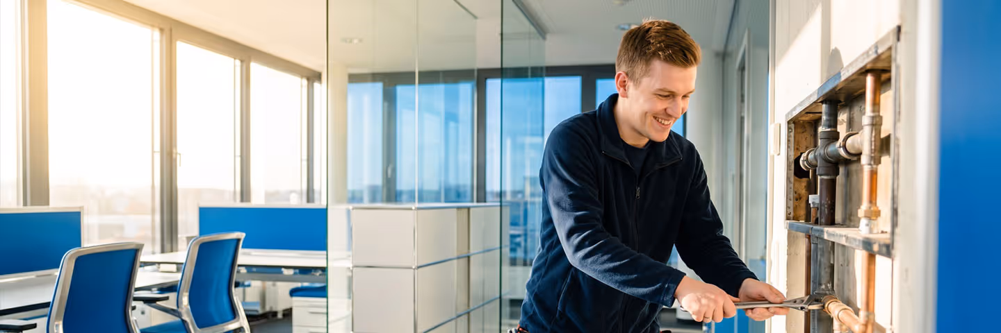 Techniker repariert Rohrleitung in modernem Büro mit großen Fenstern und blauen Stühlen.