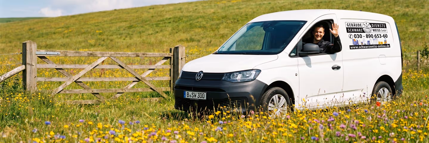 Ein weißer VW-Transporter steht in einem bunten Wildblumenfeld, Fahrer winkt aus dem Fenster.