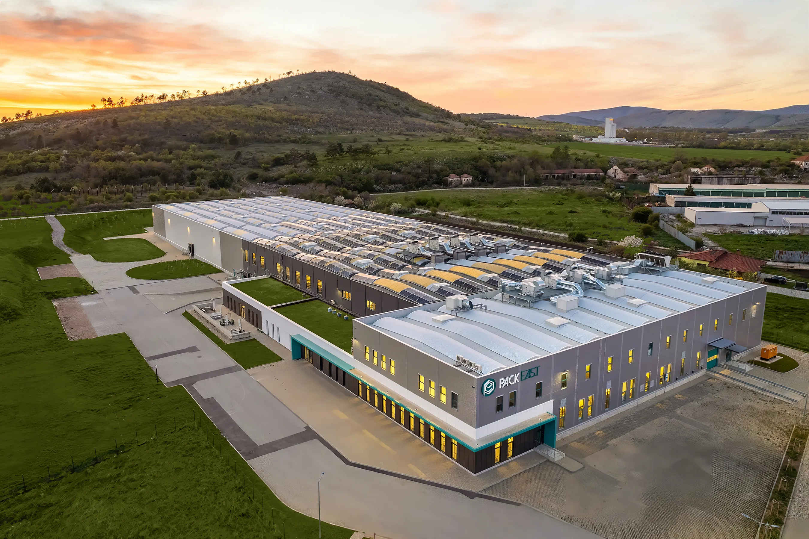 Aerial dusk view of PackEast industrial complex with illuminated barrel-vault roofs set against rolling green hills